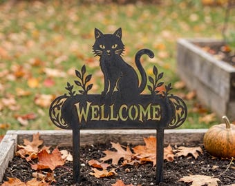 Könnte beinhalten: Ein schwarzes Metall-Gartenschild mit der Silhouette einer Katze, die auf einem rechteckigen Schild mit dem goldenen Schriftzug WELLCOME sitzt, flankiert von dekorativen Blattmotiven und zwei dünnen Stäben zur Befestigung im Boden.