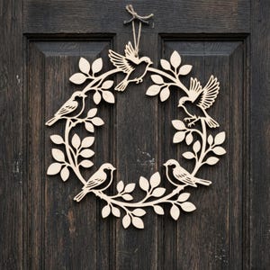 May include: A wooden wreath featuring a circular design with laser-cut birds and leaves. The birds are perched on branches, and the wreath is made of light-colored wood. It hangs on a dark wooden door.