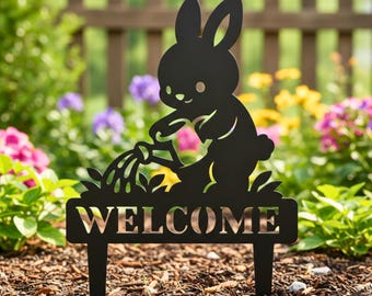 Könnte beinhalten: Schwarzer Metall-Gartenstecker mit einem Hasen, der Pflanzen gießt, mit dem Wort „WELCOME“ darunter ausgeschnitten. Der Hase hat große Ohren und einen kleinen Schwanz. Der Stecker steht in einem Garten mit bunten Blumen und einem Holzzaun im Hintergrund.