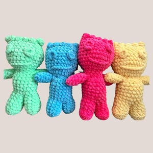 May include: Four handmade crochet bear dolls in a row, each in a different color: light green, blue, pink, and yellow. The bears have small arms and legs, and a simple face. The background is a solid light beige.