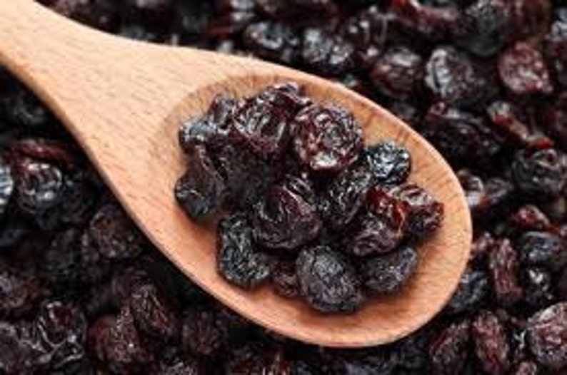 May include: A wooden spoon filled with dark brown raisins, surrounded by a pile of more raisins.