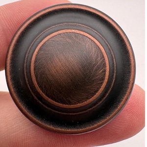 May include: A circular cabinet knob featuring a dark oil-rubbed bronze finish with copper-toned highlights on the raised center and outer rim. The hardware has a smooth, stepped profile with a brushed texture visible across the metallic surface.