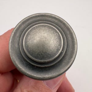 May include: A circular cabinet knob with a weathered, pewter-toned metallic finish. The hardware features a raised, rounded center dome surrounded by a flat, concentric outer ring, creating a stepped, layered profile with a matte, textured surface.