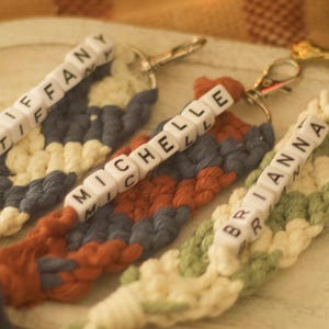 Puede incluir: Tres llaveros hechos a mano con nombres personalizados. Cada llavero presenta un diseño tejido en diferentes colores: azul y crema, óxido y azul, y verde y crema. Los nombres "TIFFANY", "MICHELLE" y "BRIANNA" están escritos en los llaveros.