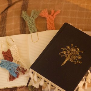 May include: An open notebook with lined pages and several handmade macrame bookmarks in various colors, including blue, brown, and white. A black journal with a gold floral design and tassel trim is also present.