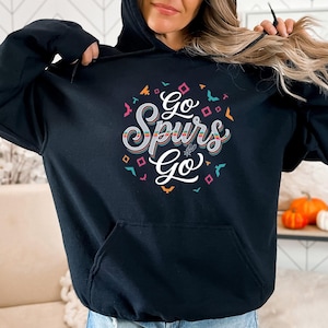 Puede incluir: Sudadera con capucha negra con el texto "Go Spurs Go" en un dise&ntilde;o colorido y en capas. El dise&ntilde;o incluye formas geom&eacute;tricas y un logotipo estilizado. La sudadera tiene un bolsillo delantero y una capucha con cord&oacute;n.