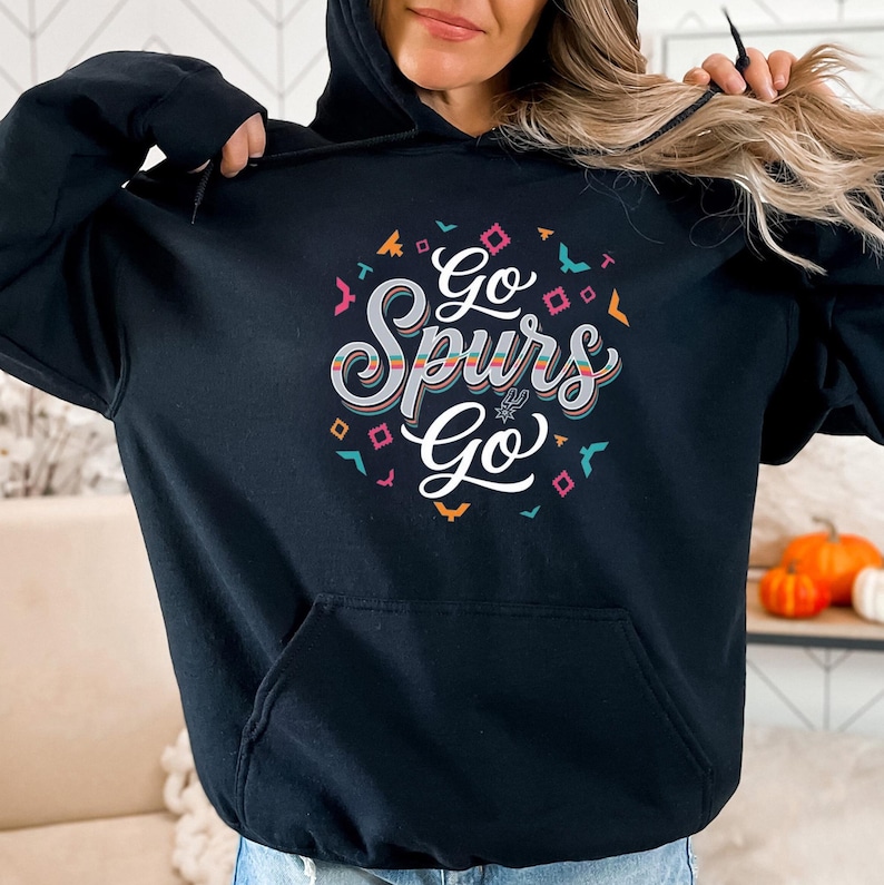 Puede incluir: Sudadera con capucha negra con el texto "Go Spurs Go" en un dise&ntilde;o colorido y en capas. El dise&ntilde;o incluye formas geom&eacute;tricas y un logotipo estilizado. La sudadera tiene un bolsillo delantero y una capucha con cord&oacute;n.