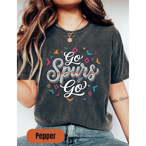 Puede incluir: Camiseta gris oscuro con el texto "Go Spurs Go" en una fuente colorida y estilizada. El dise&ntilde;o incluye una pelota de baloncesto y elementos decorativos en un patr&oacute;n circular. La camiseta es c&oacute;moda.