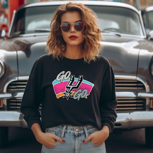 Peut inclure: Sweat-shirt noir avec l'inscription "GO SPURS GO" dans un design color&eacute;. Le graphisme comprend le logo des Spurs avec des accents roses, turquoise et orange. Le sweat-shirt est port&eacute; par une personne devant une voiture classique.