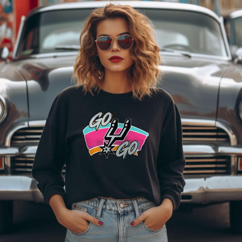 Peut inclure: Sweat-shirt noir avec l'inscription "GO SPURS GO" dans un design color&eacute;. Le graphisme comprend le logo des Spurs avec des accents roses, turquoise et orange. Le sweat-shirt est port&eacute; par une personne devant une voiture classique.