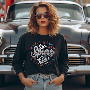 Puede incluir: Sudadera negra con el texto "Go Spurs Go" en una fuente decorativa y colorida. El dise&ntilde;o incluye formas geom&eacute;tricas e ilustraciones de p&aacute;jaros. La sudadera se combina con unos vaqueros azules.