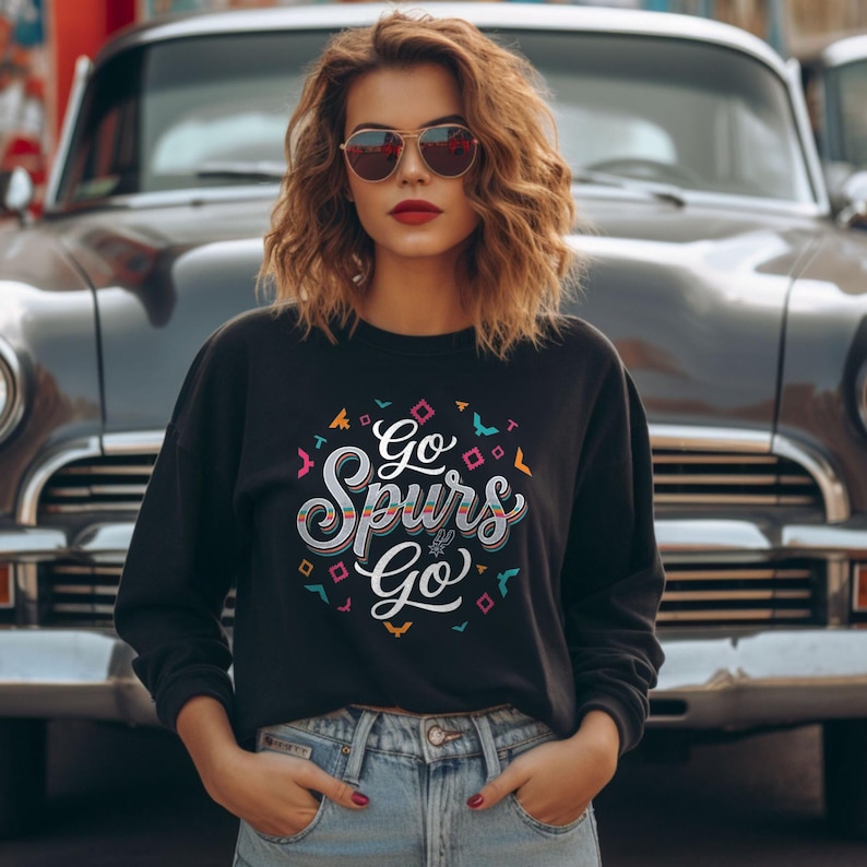 Puede incluir: Sudadera negra con el texto "Go Spurs Go" en una fuente decorativa y colorida. El dise&ntilde;o incluye formas geom&eacute;tricas e ilustraciones de p&aacute;jaros. La sudadera se combina con unos vaqueros azules.