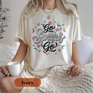 Puede incluir: Camiseta de color marfil con el texto "Go Spurs Go" en un dise&ntilde;o retro. El gr&aacute;fico incluye formas geom&eacute;tricas coloridas y detalles de p&aacute;jaros. La camiseta tiene un corte holgado y mangas cortas.
