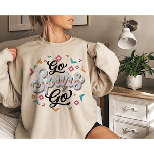 Puede incluir: Sudadera beige con el texto "Go Spurs Go" en un dise&ntilde;o estilizado y colorido. El gr&aacute;fico incluye formas geom&eacute;tricas e ilustraciones de p&aacute;jaros en varios colores. La sudadera es de estilo casual y c&oacute;modo.