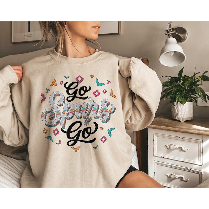 Puede incluir: Sudadera beige con el texto "Go Spurs Go" en un dise&ntilde;o estilizado y colorido. El gr&aacute;fico incluye formas geom&eacute;tricas e ilustraciones de p&aacute;jaros en varios colores. La sudadera es de estilo casual y c&oacute;modo.