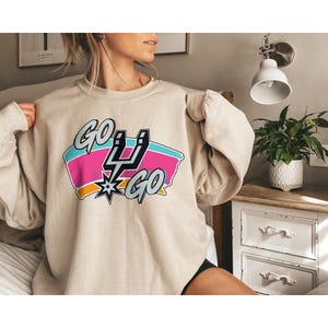 Peut inclure: Sweat-shirt beige avec un motif color&eacute; &laquo;&nbsp;GO GO&nbsp;&raquo; avec un &eacute;peron et un &eacute;clair. Le motif comprend des couleurs rose, turquoise et orange. Le sweat-shirt est de style d&eacute;contract&eacute;.