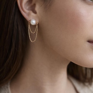 May include: A gold-toned earring featuring a large, round, white pearl stud. Two delicate gold chains dangle from the pearl, creating a layered effect. The earring is shown on an ear, highlighting its elegant design.