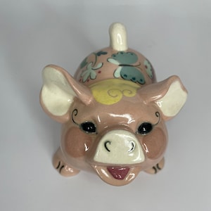 May include: A ceramic pig figurine in shades of pink, with black eyes and a smiling mouth. The pig's back is decorated with floral and butterfly designs in blue, yellow, and green. The figurine has a curled tail and is a whimsical decorative item.