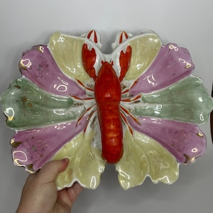 May include: A decorative ceramic serving dish in the shape of a butterfly, featuring a vibrant red lobster in the center. The dish has sections of pink, green, and yellow, with gold trim. A unique piece for serving appetizers or as a decorative accent.