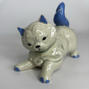 May include: A white ceramic figurine of a cat with blue ears and tail. The cat is lying on its belly with its front paws outstretched.