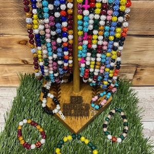 May include: A display of colorful beaded bracelets and necklaces. The jewelry features a variety of colors, including red, blue, green, and white. The bracelets are displayed on a wooden stand with the text "BB Hicks Bling".