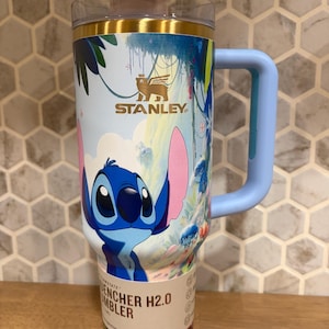 May include: A Stanley Quencher H2.0 tumbler featuring a blue Stitch character graphic, a light blue handle, a clear lid with a pink tab, and a gold-tone rim. The body displays a tropical jungle scene with colorful foliage and the Stanley logo.