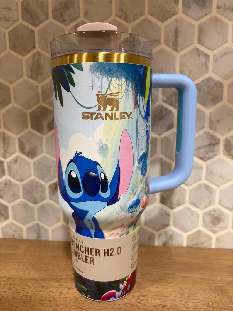 May include: A Stanley Quencher H2.0 tumbler featuring a blue Stitch character graphic, a light blue handle, a clear lid with a pink tab, and a gold-tone rim. The body displays a tropical jungle scene with colorful foliage and the Stanley logo.