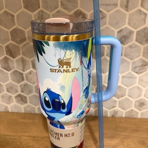 May include: A light blue Stanley Quencher H2.0 tumbler featuring a graphic of the character Stitch from Lilo and Stitch. The cup has a gold-toned rim, a clear lid with a light pink tab, a light blue handle, and a long, translucent blue straw.