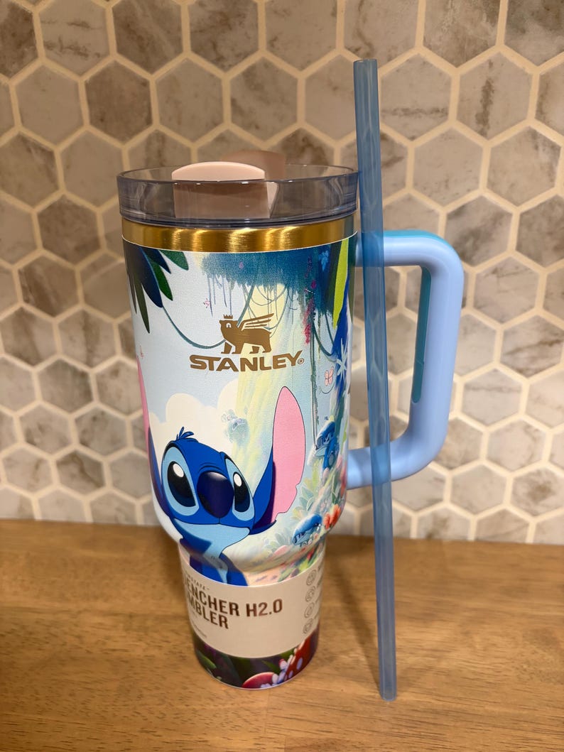 May include: A light blue Stanley Quencher H2.0 tumbler featuring a graphic of the character Stitch from Lilo and Stitch. The cup has a gold-toned rim, a clear lid with a light pink tab, a light blue handle, and a long, translucent blue straw.