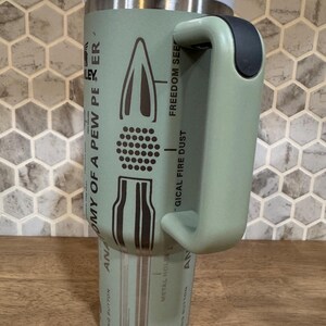 May include: A light green insulated travel mug with a white lid and black handle. The mug features a diagram of a bullet with text that reads "ANATOMY OF A PEW PER" and "FREEDOM SEE".