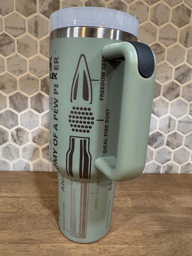 May include: A light green insulated travel mug with a white lid and black handle. The mug features a diagram of a bullet with text that reads "ANATOMY OF A PEW PER" and "FREEDOM SEE".