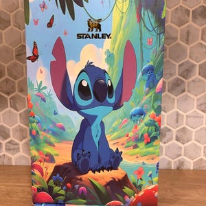 May include: A rectangular product box featuring a colorful illustration of the blue character Stitch sitting on a rock in a lush forest. The Stanley logo is printed at the top center. The scene includes vibrant flowers, butterflies, and greenery.