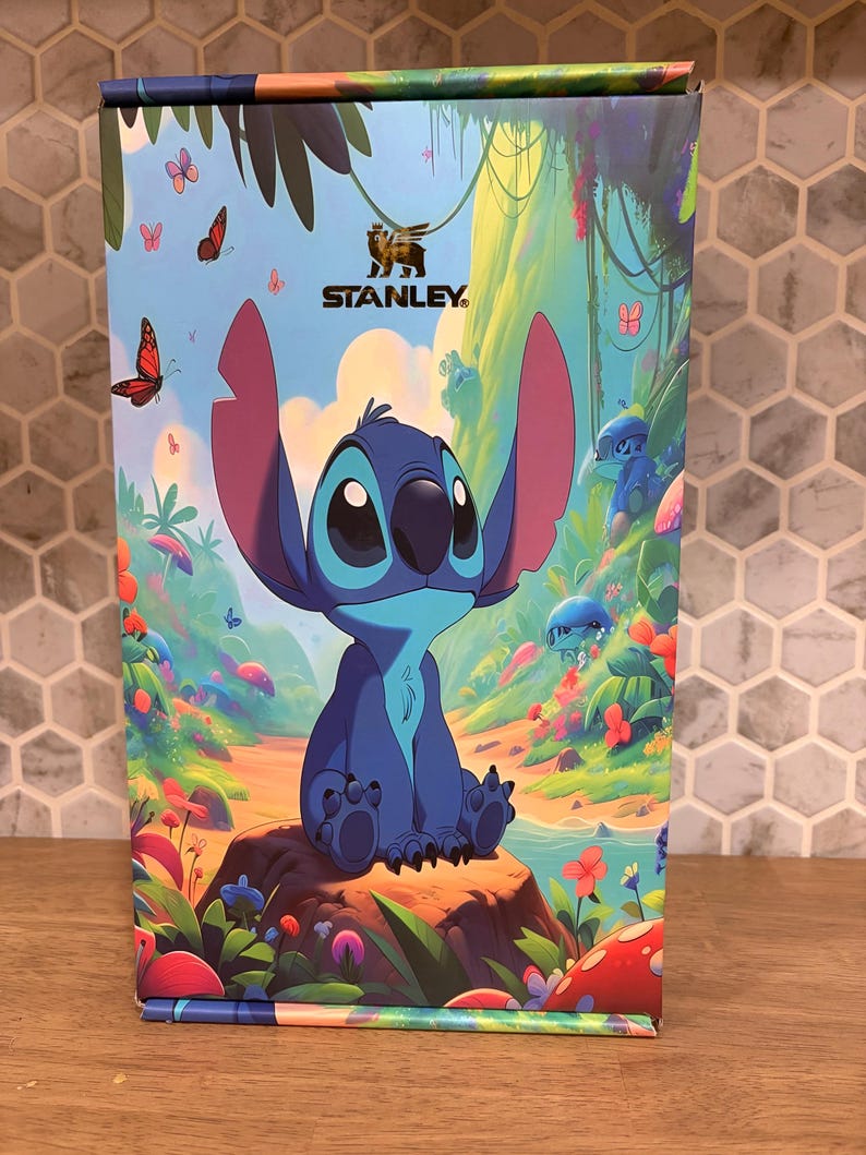 May include: A rectangular product box featuring a colorful illustration of the blue character Stitch sitting on a rock in a lush forest. The Stanley logo is printed at the top center. The scene includes vibrant flowers, butterflies, and greenery.