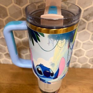 May include: A light blue insulated tumbler with a handle and a clear lid featuring a pink sliding closure. The exterior displays a colorful graphic of the character Stitch from Lilo and Stitch set against a tropical jungle background with green leaves.