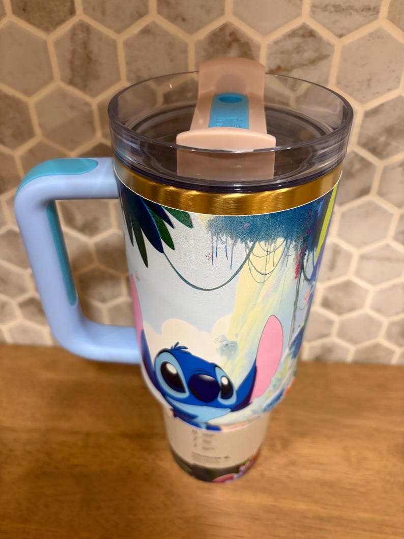 May include: A light blue insulated tumbler with a handle and a clear lid featuring a pink sliding closure. The exterior displays a colorful graphic of the character Stitch from Lilo and Stitch set against a tropical jungle background with green leaves.