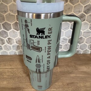 May include: A mint green Stanley travel mug with a handle and a white lid. The mug features the Stanley logo and the words "ANATOMY OF A PEW PEER" with illustrations of bullets and other gun-related imagery.