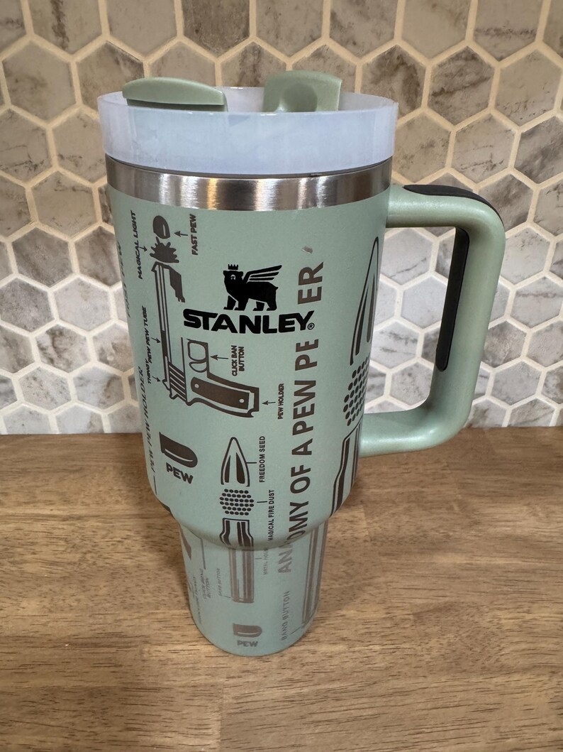 May include: A mint green Stanley travel mug with a handle and a white lid. The mug features the Stanley logo and the words "ANATOMY OF A PEW PEER" with illustrations of bullets and other gun-related imagery.