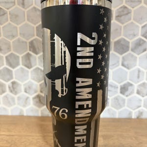 May include: A matte black stainless steel tumbler featuring a laser-engraved design of a stylized skull, the text 2ND AMENDMENT, the number 76, and a vertical American flag motif. The tumbler has a black plastic lid and a tapered cylindrical body.
