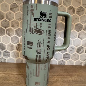 May include: A Stanley brand insulated tumbler in a muted green color, featuring a handle and a screw-on lid. The tumbler is adorned with illustrations and text, including the words "ANATOMY OF A PEW PEER" and "PEW". The Stanley logo is also visible.