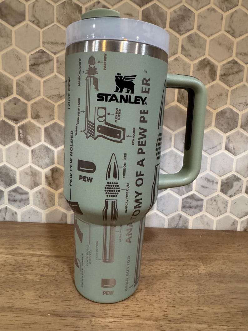 May include: A Stanley brand insulated tumbler in a muted green color, featuring a handle and a screw-on lid. The tumbler is adorned with illustrations and text, including the words "ANATOMY OF A PEW PEER" and "PEW". The Stanley logo is also visible.