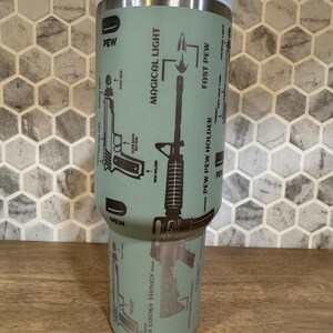 May include: A light green tumbler with a white lid features diagrams of firearms and the phrases "PEW," "MAGICAL LIGHT," and "FAST PEW." The tumbler has a stainless steel rim and a design that includes the text "AIM LOOKY THINGY."