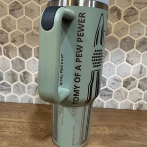 May include: A light green insulated tumbler with a handle, a white lid, and black accents. The text "ANATOMY OF A PEW PEWER" and other phrases are printed on the side. The tumbler is on a wooden surface.