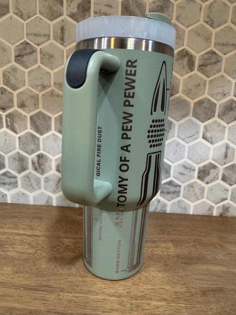 May include: A light green insulated tumbler with a handle, a white lid, and black accents. The text "ANATOMY OF A PEW PEWER" and other phrases are printed on the side. The tumbler is on a wooden surface.