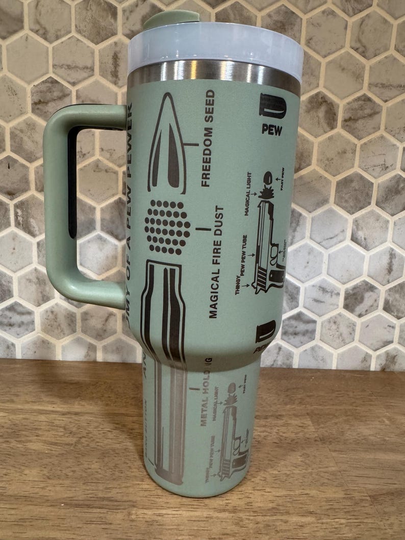 May include: A light green insulated tumbler with a handle and a light gray lid. The tumbler features illustrations and text including "Freedom Seed", "Magical Fire Dust", and "Pew".