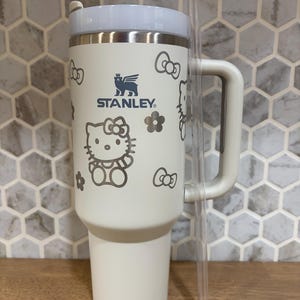 May include: A cream-colored Stanley tumbler featuring a Hello Kitty pattern with bows and flowers. The insulated cup has a sturdy side handle, a white lid, and a clear plastic straw. The Stanley logo is printed in dark blue on the front surface.