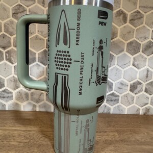 May include: A light green insulated travel mug with a handle and a light blue lid. The mug features illustrations of bullets, guns, and text including "Freedom Seed" and "Magical Fire Dust."