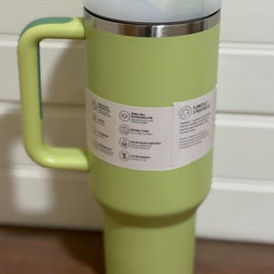 K&ouml;nnte beinhalten: Ein hellgr&uuml;ner isolierter Becher mit Griff und Deckel. Der Becher hat einen Edelstahlrand und hellgr&uuml;ne Griff- und Deckelakzente. Ein wei&szlig;es Etikett umschlie&szlig;t den Becher mit Text und Symbolen.