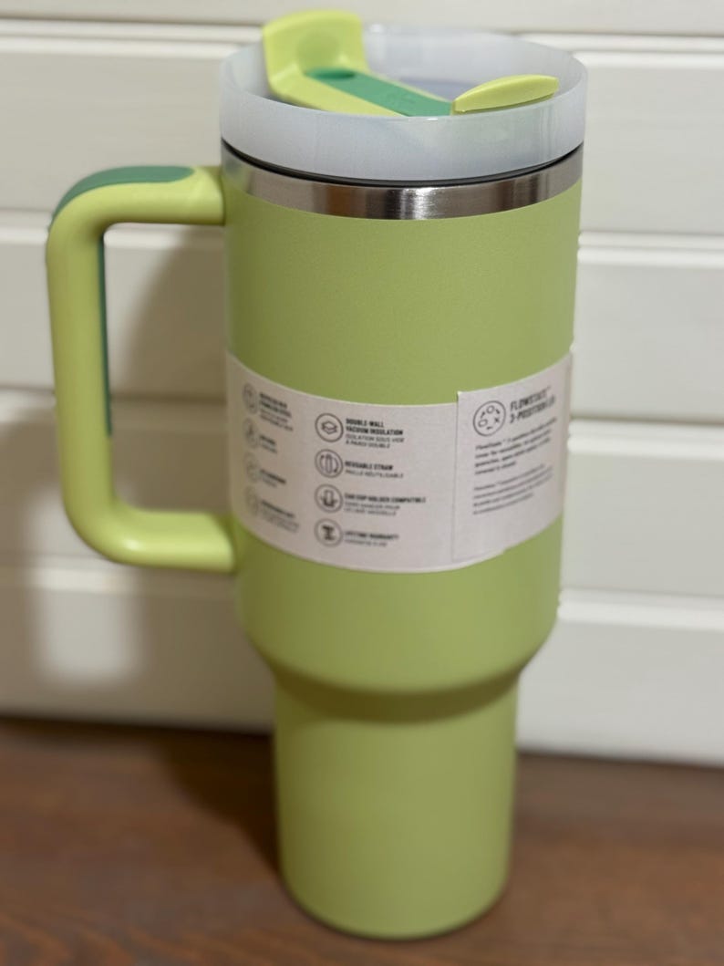 K&ouml;nnte beinhalten: Ein hellgr&uuml;ner isolierter Becher mit Griff und Deckel. Der Becher hat einen Edelstahlrand und hellgr&uuml;ne Griff- und Deckelakzente. Ein wei&szlig;es Etikett umschlie&szlig;t den Becher mit Text und Symbolen.
