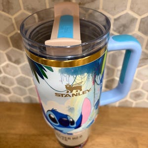 May include: A Stanley brand tumbler featuring a light blue handle, a clear lid with a light pink and blue sliding closure, and a wrap-around graphic of the character Stitch from Lilo and Stitch set against a tropical background with palm leaves.