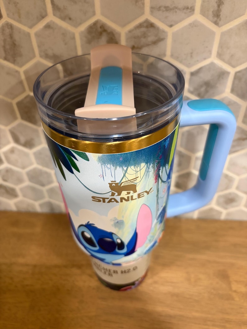 May include: A Stanley brand tumbler featuring a light blue handle, a clear lid with a light pink and blue sliding closure, and a wrap-around graphic of the character Stitch from Lilo and Stitch set against a tropical background with palm leaves.